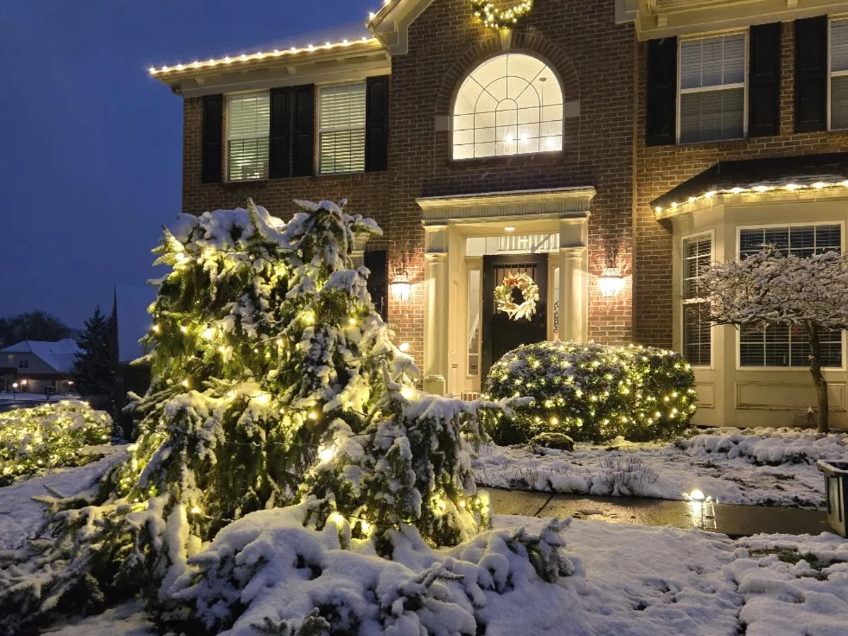 Snow-covered evergreen wrapped in warm lights beside a front entry decorated with a Christmas wreath