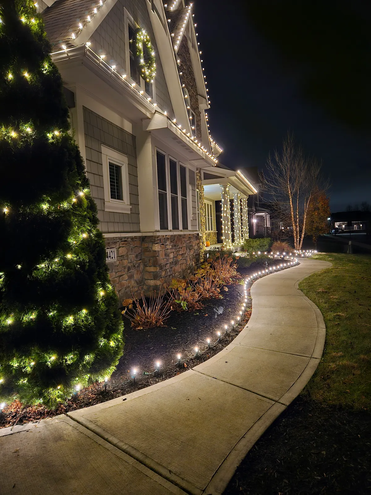 Home with warm-white Christmas lights on the eaves, a lit wreath on the gable, and wrapped porch columns