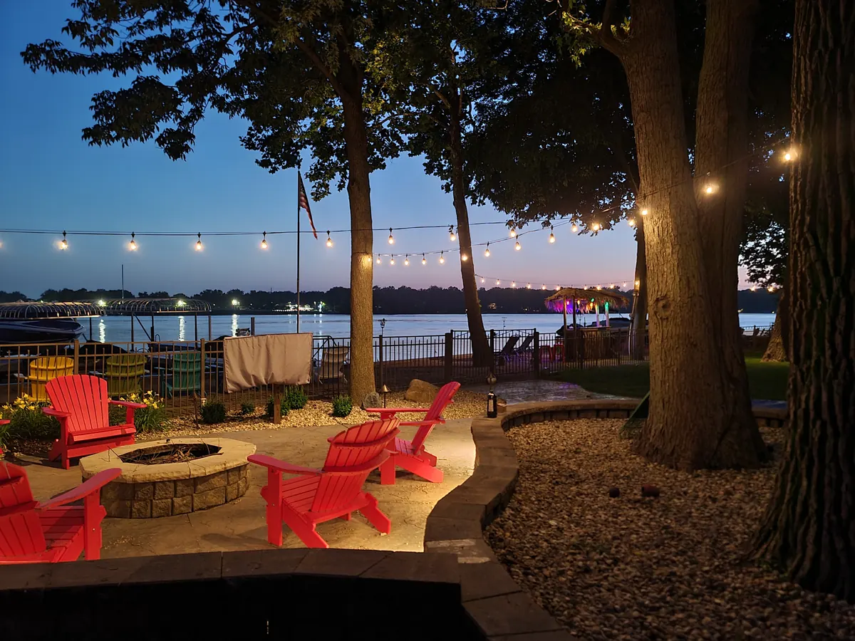 Waterfront pavilion at dusk with overhead string lights and red Adirondack chairs