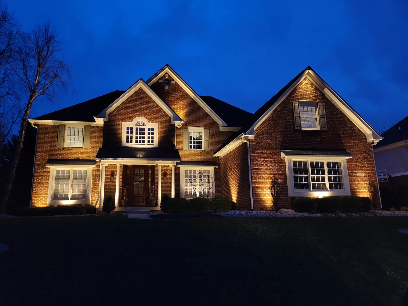 Warm-white architectural uplighting on a brick colonial home at dusk — the same fixtures running in everyday accent mode