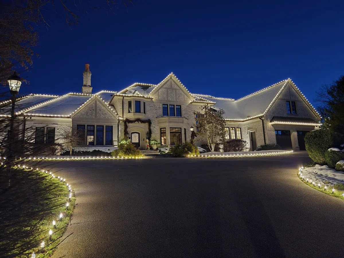 Warm-white Christmas roofline lighting and lit driveway borders on a snowy stone home at dusk
